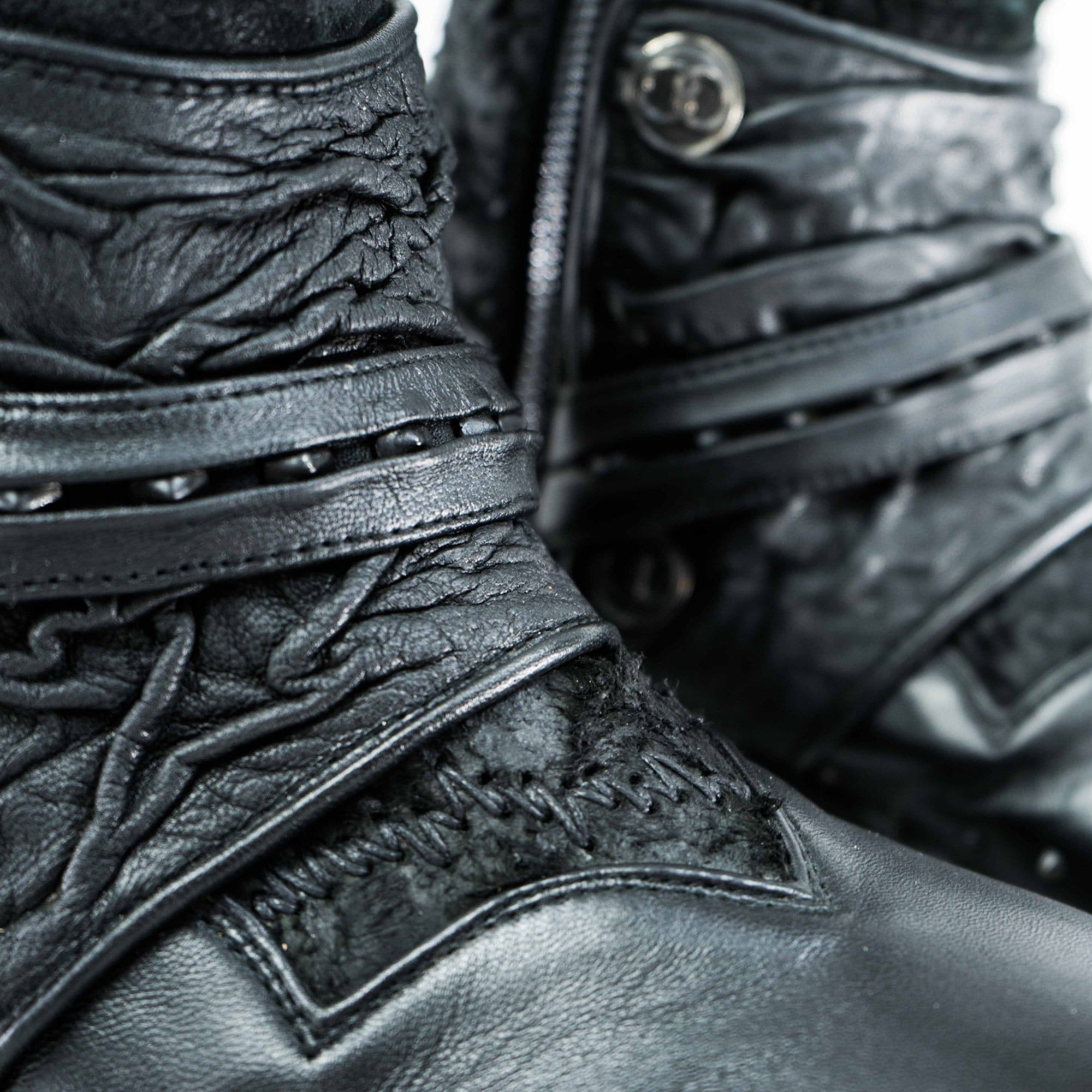 Close-up of a pair of MAGMA Wasteland Studded Leather Ankle Combat Boots by Kaya Movement, showcasing their textured, wrinkled detailing and multiple straps including a studded strap and one with a button closure. The intricate design and stitching are prominently featured.